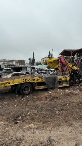 The video shows a scene of destruction and debris. A yellow tow truck with a crane is present, apparently assisting in removing a mangled vehicle. The surrounding area is strewn with debris, including broken trees, rubble, and twisted metal. A large hole is visible in the ground, suggesting a significant impact or explosion. There are a few buildings in the background that show signs of damage. Two people can be seen standing near the edge of the hole. The overall impression is one of a site affected by a catastrophic event.