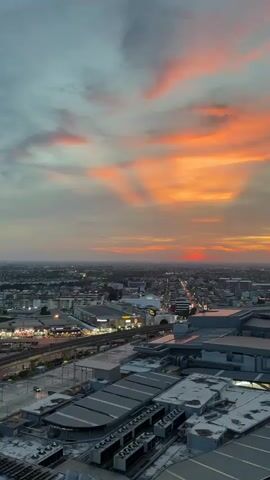 An aerial, vertical shot shows a cityscape at dusk. The sky is filled with dramatic clouds painted in hues of orange, pink, and gray, with visible sunbeams radiating downwards. Below, the urban landscape stretches out, dotted with lights from buildings and streets. In the foreground, the rooftops of modern structures, possibly commercial or industrial buildings with intricate ventilation systems, are visible. A railway line with a train passing by is discernible to the left of the frame, adding a sense of movement to the otherwise static scene. The overall impression is one of an expansive city bathed in the warm glow of a sunset.