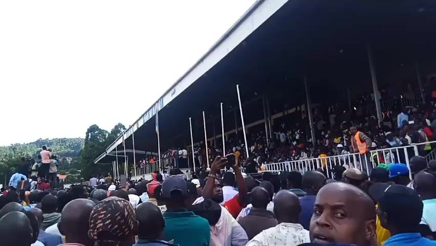 Here's a concise description of the video:

The video shows a large crowd of people gathered outside a stadium. The crowd is dense, and many people are looking up towards the stadium's seating area, which appears to be filled with spectators. Some individuals are recording with their phones, and a few people are standing on raised objects to get a better view. The sky is partly cloudy. The stadium itself has a long, covered seating area supported by pillars. The environment suggests a sporting event or a rally.