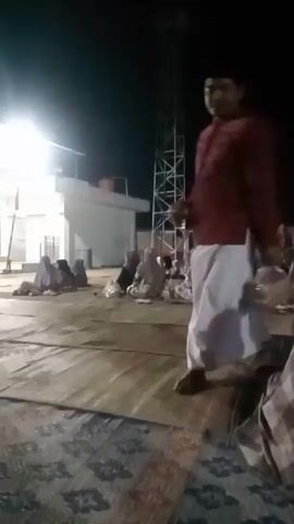 A group of people are gathered outdoors at night. A man in a red shirt and white pants is standing and appears to be talking or presenting something. Several other people are sitting on mats on the ground, some in the background appear to be women wearing hijabs. There is artificial lighting in the scene, possibly from a nearby building and a tall metal structure that might be a tower. The ground is covered with mats and possibly rugs. The overall atmosphere suggests a communal gathering or event.