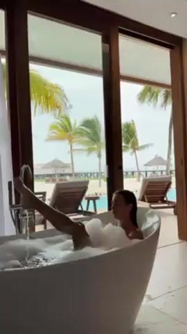 A woman is in a white bathtub filled with bubbly water, with one leg raised straight up in the air. The tub is positioned in front of a large window overlooking a tropical scene with palm trees, a beach, lounge chairs, and a pool. The sky is overcast. The bathroom has wood-framed sliding doors and a modern faucet next to the tub.