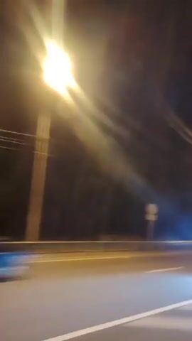 The video shows a nighttime view from a moving vehicle. The scene is primarily dark with streaks of light from streetlights and passing vehicles. The road is visible with white lines. On the side of the road, there are trees and some structures, including what appears to be a bus stop shelter in one shot and a traffic light in another. A car with its headlights on is seen passing by. The overall impression is a high-speed drive on a road at night, possibly a highway or a major road.
