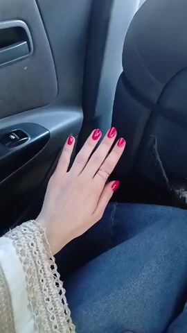 The video shows the left hand of a person inside a car, moving slightly. The person is wearing a ring on their ring finger and has bright red nail polish. The sleeve of a white, lace-trimmed garment is visible on the person's wrist. The person is wearing blue jeans. The car interior is dark gray, with a door panel visible on the left and a seat back on the right.