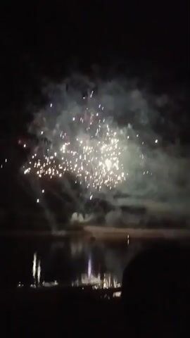 A fireworks display is happening over a body of water at night. The fireworks are white and gold, creating a shimmering effect against the dark sky. There are also some faint blue and red lights reflected in the water. The silhouette of people watching the display can be seen in the foreground.