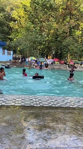 A group of people, mostly children, are enjoying a swimming pool surrounded by lush green trees. The water is a vibrant blue, and some individuals are using inflatable rings. In the background, tents are set up, suggesting a recreational area. The foreground shows a wet concrete path leading to the pool. One person, partially visible, is walking away from the camera, while another person is crawling out of the pool. The scene appears to be a lively and casual outdoor gathering.