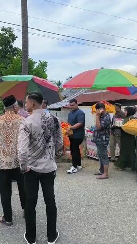A group of people are gathered outdoors, some carrying parcels and gifts. One person is wearing a traditional headdress, suggesting a celebratory event, possibly a wedding. A multi-colored umbrella provides shade. A motorcycle is visible in the foreground. The environment appears to be a street with houses in the background, indicating a residential area. The overall atmosphere suggests a community gathering.