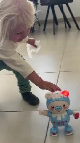 A toy pig dances on a tiled floor. A person's legs, clad in gray socks and green pants, are visible in the upper left corner of the frame, moving out of frame. The toy pig is blue and white, with pink cheeks and red lights on its head and hands. It moves its arms and legs and appears to be "dancing". The background consists of a tiled floor and the legs of two chairs.