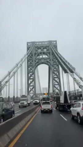The video shows a gray suspension bridge from the perspective of a car moving along a roadway. Several lanes of traffic are visible, with a mix of cars and trucks traveling in the same direction. The sky is overcast and gray. The camera seems to be mounted inside a vehicle, looking forward. A median barrier separates the car's lane from opposing traffic.
