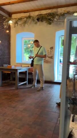 A man wearing a light blue t-shirt and beige pants stands in a room, looking at his phone. He is wearing a black bag across his chest. The room has exposed brick walls, wooden beams on the ceiling, and a large window. There are several tables and chairs in the room, as well as decorative lights. The floor is tiled. There are plants hanging from the ceiling and a mural on one of the walls.