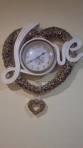 The media file shows a close-up, eye-level, static shot of a decorative heart-shaped clock. The clock face is circular and positioned in the center of the heart, with Roman numerals marking the hours and two hands indicating the time. The word "Love" is elegantly written in white script and integrated into the heart shape, wrapping around the clock face. The heart itself is adorned with a textured, intricate pattern of what appear to be small roses or floral elements, finished in a bronze or antique gold color. A decorative heart-shaped pendulum hangs from the bottom of the clock. The clock is mounted on a plain, light-colored wall, likely indoors. No people are visible in the media file. The lighting appears to be artificial, creating some subtle shadows and highlighting the textures of the clock. The overall impression is one of romantic or decorative intent. There is a small inscription on the clock face that reads "Le Mans." The time on the clock appears to be approximately 1:50. Given the context of Chernomorskiy, Russia, this is likely an indoor shot within a home or establishment.