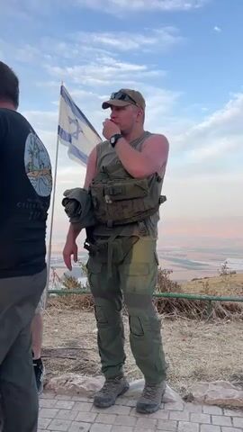 The video shows a man wearing a green military vest and pants standing outdoors, with an Israeli flag waving in the background. He is putting something red in his mouth, possibly a cherry or small fruit. He is surrounded by other people, one of whom is standing beside him with his arms crossed. The background features a view of a valley or agricultural area. The lighting suggests it is late afternoon or early evening.