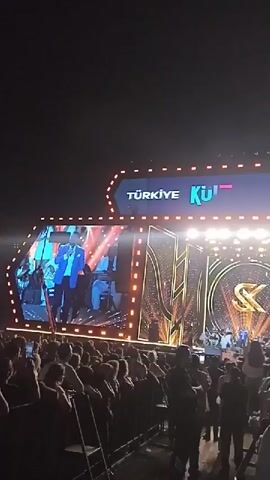 The video shows a large outdoor concert venue at night, with a stage set up for a performance. The stage is brightly lit with a large screen displaying the performers on stage. The stage has decorative elements with an 'SK' logo visible. A large crowd of people are standing in front of the stage, watching the performance and there are metal barriers keeping them back. Above the stage, a banner reads “Türkiye Kültür Yolu Festivali”.  The camera is positioned within the crowd and appears to be handheld, with slight movements. Overall, the video captures the atmosphere of a live music event.