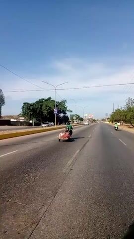 The video shows a view from a moving vehicle on a multi-lane highway. A motorcycle with a sidecar attached, carrying a load of what appears to be red meat, is traveling in the same direction in the right lane. There are other vehicles on the road including cars, a truck, and another motorcycle further ahead. The sky is blue with scattered clouds. Streetlights line the sides of the road. Trees and some buildings are visible in the background. The road surface is asphalt with white lane markings.