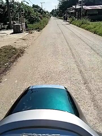 The video depicts a first-person perspective from a moving scooter driving down a paved road.  The scooter has a glossy teal and silver body visible in the foreground. The road is bordered on either side by vegetation, telephone poles, and occasional buildings. The sky is blue with scattered clouds. There are two pedestrians visible in the distance on the right side of the road. Another scooter is traveling down the road in the same direction as the first one. The sound of wind noise is prominent.