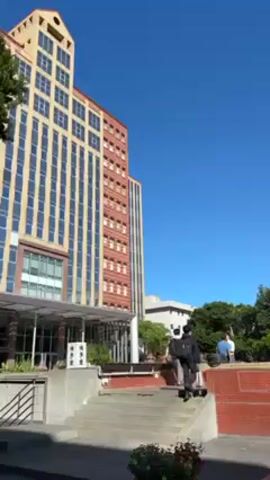 The video shows an outdoor scene under a clear blue sky. The camera is positioned low and looks upwards, initially focusing on a tall, multi-story building with a facade composed of alternating vertical bands of light beige and reddish-brown. The building has numerous windows. In front of the building, there's a paved area with a set of stairs leading up to a slightly elevated platform. On this platform, a few people are sitting and standing. One person is walking across the platform. The camera then pans right to show a second, white-colored building behind a row of neatly trimmed trees. The camera briefly focuses on a blue and white sign next to a ramp leading downwards. The sign includes directional information along with a wheelchair symbol, and text in a non-English language.