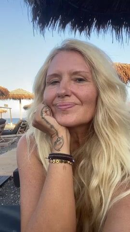 A woman with blonde hair smiles and rests her chin on her hand. She is wearing bracelets and has a tattoo on her hand. In the background, there are beach chairs and umbrellas on a pebble beach. The sky is clear and blue.