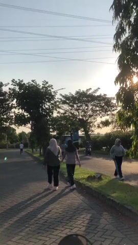 The media file depicts an outdoor scene at dusk in Kludan, East Java. The sun is low in the sky, casting long shadows and creating a warm, golden light, suggesting it is either sunrise or sunset. The weather appears clear with a bright sky.

The primary subjects are two individuals, likely young women, walking away from the camera on a paved path. They are dressed in casual athletic wear, with one wearing a headscarf. Behind them, other people are visible in the distance, also appearing to be engaged in outdoor activities like walking or jogging. A few trees line the path, with dense foliage. In the background, there are hints of more vegetation and possibly some structures, though they are indistinct. Overhead, power lines are visible against the sky, indicating an urban or semi-urban environment.

The activity taking place is leisurely walking or exercising outdoors, likely a daily routine given the presence of multiple individuals. The overall mood conveyed is peaceful and serene, characterized by the calm atmosphere and the warm lighting. There is no visible text in the media file.