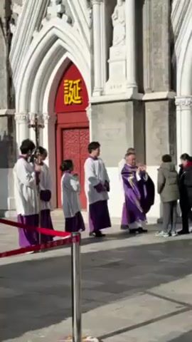 A small procession of people wearing white and purple robes and a priest in a purple robe stand outside a church with large red doors. The church is white with a statue on one side of the entrance. A cross is being held by a person in the group. People are walking past them. The priest appears to be distributing something, maybe money or a small card, to people passing by. A red rope attached to a metal stanchion is visible in the foreground.