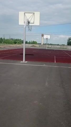 The video shows a maroon outdoor basketball court. The court is empty and has two basketball hoops at either end. The hoops have white backboards and silver poles. The court is surrounded by a gray asphalt surface. In the background, there is a water tower and some greenery. The sky is partly cloudy.