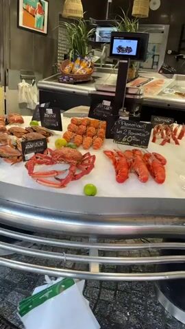 The video pans across a seafood display, likely in a market or restaurant. The display is covered in ice and features a variety of fresh seafood, including crabs, lobsters, and what appear to be fish fillets. Lemons and limes are interspersed among the seafood for decoration and possibly flavor. In the background, there are signs with prices and descriptions, a basket of bread or pastries, and a digital screen displaying information. The overall impression is one of a well-stocked and appealing seafood presentation.