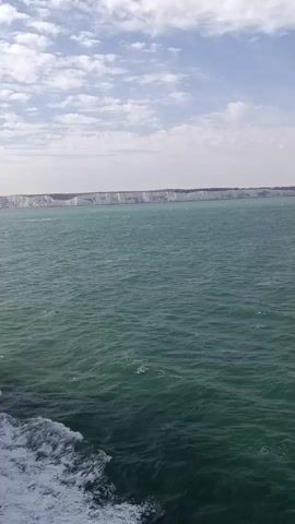 A wide shot captures a vast expanse of choppy, green water under a partly cloudy sky. In the distance, a long, white cliff face stretches across the horizon, its rugged edge disappearing into a hazy sky. The foreground shows the foamy wake of a vessel, indicating movement through the water. The overall impression is one of a journey across open water, with a dramatic coastline in view.