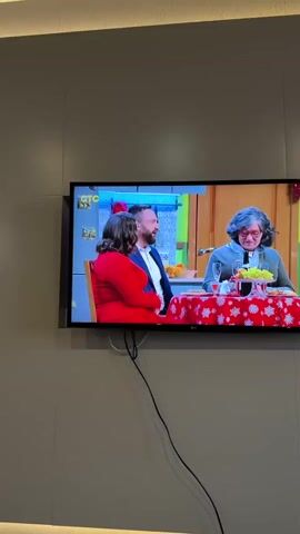 A television is mounted on a beige wall. The television is showing a scene with three people sitting at a table decorated for the holidays. The table has a red tablecloth with white snowflakes, and there are glasses and fruit on it. One of the people is wearing a red outfit. The other man is wearing a suit, and the other person appears to be in costume, wearing a gray wig, glasses, and clothing that makes them look like an older woman. The woman in the red outfit speaks, and the camera zooms in on her face. Then, the camera zooms in on the face of the person in the gray wig. The TV is an LG model. There are two black cords hanging beneath the TV. A portion of a light fixture can be seen at the top of the wall. The logo for the TV channel CTC is visible in the upper-left corner of the TV screen.
