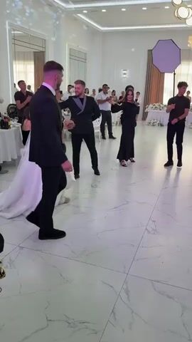 A wedding party is dancing in a large, bright hall. The bride and groom are in the center, dancing with other guests around them. The bride is wearing a white wedding dress with a long veil, and the groom is wearing a black suit. There are several people standing and watching the dancers, some of whom are holding their phones up to take pictures or videos. The hall has a high ceiling with decorative lighting fixtures, and the floor is made of white marble with grey veins. The walls are white, and there are large windows with sheer curtains. There are tables with floral arrangements in the background.