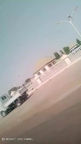 The video shows a moving vehicle driving on a paved road near what appears to be a dirt hillside. Other vehicles, including a small white truck, are visible on the road. Buildings and power lines are also visible in the background. The video seems to be recorded from within another moving vehicle. The camera angle is skewed, and there is some shakiness. A logo "SHOT ON P6S_" is displayed on the screen. There's some talking in an African language in the background.