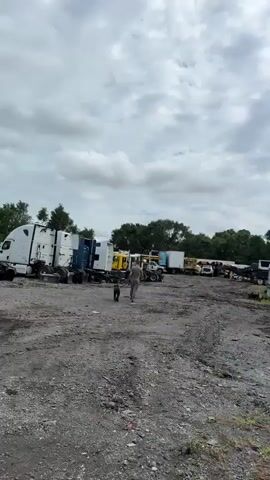 The video opens with a wide shot of a junkyard with several trucks. The camera pans to the right, showing rows of semi-trucks parked in an outdoor lot. Many of the trucks appear to be damaged or disassembled, with hoods up and parts missing. Some of the trucks are black, while others are white. In the foreground, there is a pile of scrap metal and debris. A person is visible walking a dog in the distance on the left side of the frame. The sky is overcast. The camera then focuses on a group of black semi-trucks parked closely together, with their hoods open. Following this, the camera shows the rear of a white semi-truck, highlighting its wheels and mud flaps. The video concludes with a shot of the side of the same white semi-truck, showing its wheels and the surrounding gravel lot. The environment appears to be an industrial area or scrap yard.