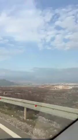The video shows a scene from a moving car. The car passes by a guardrail on the side of the road, with a vast landscape visible in the background. The sky is blue with patchy clouds. Further away, there's a mix of trees, barren land, and a town. At one point, the car passes by a small, dilapidated building covered in graffiti. Upbeat music is playing in the background.