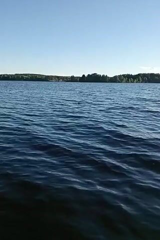 A first-person perspective video shows feet dangling over the edge of what appears to be a dock or boat. The water is dark blue with ripples, and the sky is clear blue. In some shots, the legs are visible, bent at the knees, and the person is wearing light-colored shorts. There are trees and buildings visible in the distance on the far shore of the lake. The feet are bare. The camera pans slightly, showing the movement of the water and the legs dipping into the water at one point.