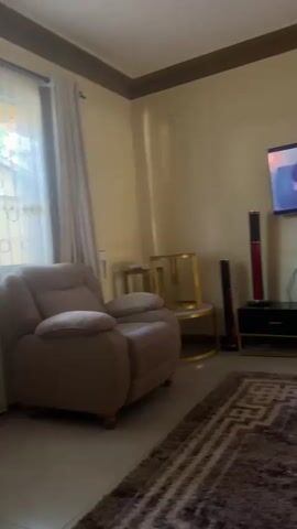 A person is sitting in a brown recliner chair in a living room, watching television. The television is mounted on the wall above a black entertainment center. There are two tall, red and black speakers on either side of the entertainment center. A patterned rug covers the floor in front of the entertainment center. The walls are painted a light yellow color and there is a brown decorative trim along the top of the walls. A window with a light-colored curtain is visible on the left side of the screen.