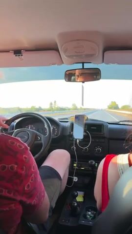 The video shows the interior of a vehicle traveling down a highway at sunset. A man in a red patterned shirt and shorts is driving, with his left hand on the steering wheel. A woman in a red top is in the passenger seat. A navigation app is displayed on a phone mounted to the dashboard. In the backseat, a woman in a striped shirt is taking a selfie, with luggage visible behind her. The exterior environment appears to be a flat, open landscape with trees lining the highway. The lighting suggests it is late afternoon or early evening.