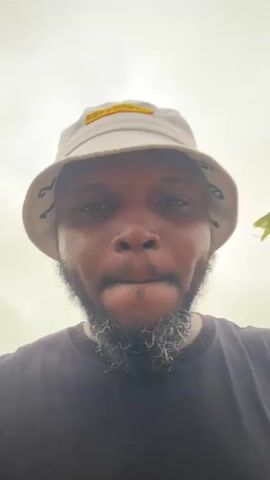 The video shows a close-up, low-angle shot of a Black man wearing a bucket hat and a dark t-shirt. He has a beard with some gray hairs. The hat is light-colored with black lettering around the brim and a yellow patch on the front. He is looking up and to the side, then turns his head to face the camera. At one point, he raises his right hand towards his face, obscuring it slightly. He appears to be outdoors, with a bright, overcast sky in the background. There is a plant with red leaves visible to the right of his head at different points. His left wrist has an orange watchband.