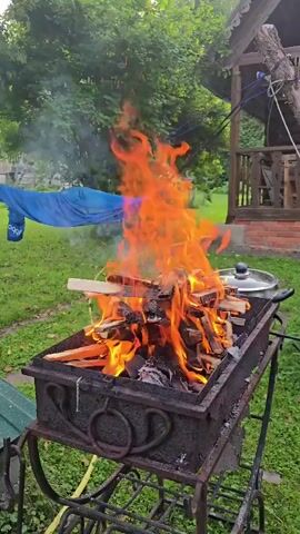 A fire burns in a metal grill outdoors. The grill is filled with wood logs, some of which are on fire, producing large flames. A lid rests on the edge of the grill. In the background, a blue tarp hangs from a clothesline between two trees, and a wooden structure is visible. The ground is covered in green grass.