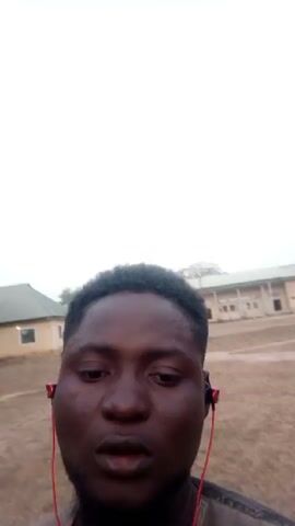 The video features a close-up of a dark-skinned man, who appears to be outside in a somewhat hazy or overcast environment. He has short, curly hair and a beard. He is wearing red earphones and a grey or brown shirt. In the background, there are buildings and a dusty ground area. He sticks out his tongue briefly, puckers his lips, and then shields his eyes with his hand.
