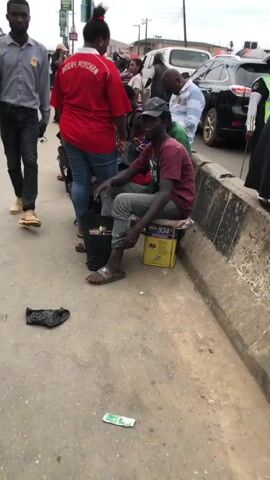 The video shows a street scene with several people. In the foreground, a man walks past the camera, wearing a collared shirt, jeans, and light-colored shoes. A dark plastic bag lies on the ground in front of him. To his right, a woman in a red shirt stands near a motorcycle. A man wearing a maroon shirt and a dark cap is sitting on a yellow box, looking towards the camera. Behind him, more people are visible, some sitting on a raised concrete curb. They appear to be in a market or commercial area, with stalls and goods lining the street. There are also vehicles, including a white van and a yellow auto-rickshaw, in the background. The environment looks like a busy urban street with a dusty surface and overcast sky.
