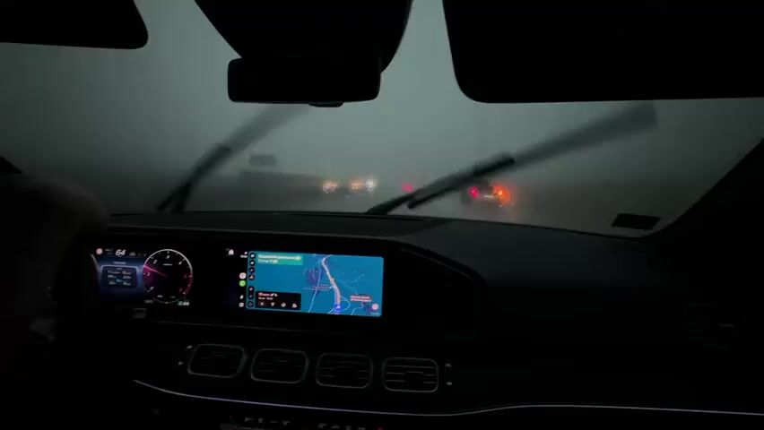 This is a point-of-view shot from inside a car during a rainstorm. The windshield is wet and the wipers are active, clearing visibility. The dashboard is lit, showing a digital instrument cluster on the left and a navigation screen with a map on the right. A hand is visible on the steering wheel. Outside, traffic lights and headlights of other vehicles are visible through the rain, indicating a road environment with other cars. The overall atmosphere is dark and hazy due to the weather conditions.