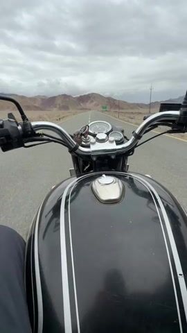 A first-person perspective video shows a rider on a black motorcycle with white pinstripes. The rider's dark pants are visible at the bottom of the frame. The motorcycle's handlebars, speedometer, and fuel tank are in the foreground. The motorcycle is on an asphalt road, which stretches into the distance, flanked by arid, mountainous terrain under a cloudy sky. A green sign is visible in the distance on the right side of the road. The motorcycle is moving forward.