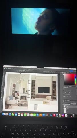 The video displays a dimly lit indoor scene, likely at night, with a laptop open on a surface and a video projected onto a wall above it. The laptop is a MacBook Pro, indicated by its design and the macOS interface, and features a keyboard with both Latin and Cyrillic characters.

The laptop screen is actively displaying Adobe Photoshop 2023. Within Photoshop, two interior design mock-ups are visible: one depicting a cozy living room with a fireplace and stylish furniture, and another showcasing a modern wall unit with a built-in television, shelving, and cabinets. A mouse cursor is positioned on the Photoshop canvas, suggesting ongoing work or editing. Visible text on the Photoshop interface includes "Adobe Photoshop 2023", "Слои" (Layers), and "Без имени" (Untitled), indicating a Russian-speaking user. The macOS Dock is also visible, showing icons for numerous applications, including various Adobe Creative Suite programs (Photoshop, Illustrator, Premiere Pro, After Effects, InDesign), communication apps (Telegram, WhatsApp, Zoom), and productivity tools.

Above the laptop, a video is projected, showing a close-up of a young man's face, illuminated by a soft blue light. He is looking upwards with a serious or contemplative expression. The projected video and the laptop screen are the primary light sources in the otherwise dark environment.