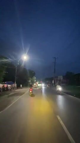 The video shows a dark street scene at dusk or dawn. Several vehicles, including motorcycles, are driving on the road, with some headlights visible. The street appears wet, suggesting recent rain. Streetlights are illuminated, casting light on the surroundings. A few people are visible on the side of the road, and some buildings or shops line the street. The sky is a deep blue, indicative of low light conditions.