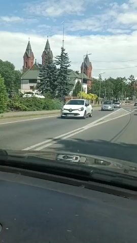 This media file captures a view from inside a vehicle driving on a street in Ostrów Mazowiecka, Poland. The foreground is dominated by the dashboard of the car, suggesting the perspective of the driver or a passenger. The road ahead is paved, with clear lane markings and a pedestrian crossing visible. Several cars are on the road, including a white hatchback in the left lane and a silver minivan in the right lane.

In the background, a striking architectural feature stands out: a red-brick church with multiple spires, characteristic of Polish religious architecture. The church appears to be a prominent landmark in the area. Adjacent to the church are residential buildings and lush green trees, suggesting a mixed urban and suburban environment. The sky is blue with scattered white clouds, indicating fair weather during the daytime. The overall mood is serene and observational, as the vehicle moves through the town. No explicit text is visible in the media file, other than license plates on the vehicles which are too indistinct to read.