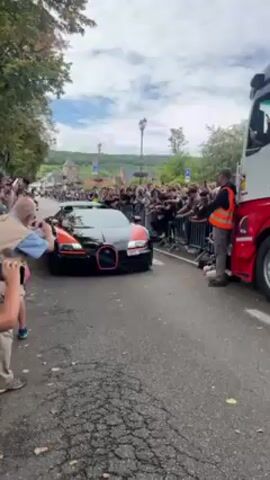 A black and orange Bugatti Veyron car, with two men inside, rolls along an asphalt street lined by a crowd of onlookers. Most in the crowd are photographing or filming the car with cameras or phones.  The road is cracked and dry. A large truck is parked on the right side of the road, and a man in a high-visibility vest stands near it. Buildings and trees are visible in the background. The sky is partly cloudy.