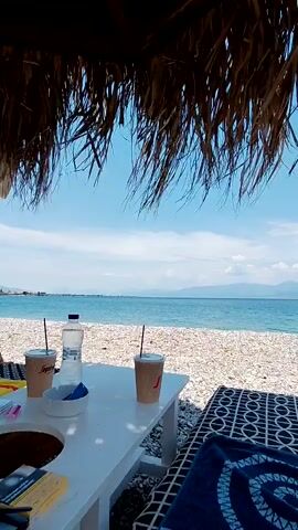 This is a first-person perspective video taken from under a thatched roof looking out at a pebble beach and the ocean. In the foreground, a white table holds two paper coffee cups with straws, a bottle of water, and an ashtray. To the right of the table is a patterned lounge chair with a blue towel on it. The ocean stretches to the horizon, with a clear blue sky and some clouds above. Mountains are visible in the distance on the left side of the frame. The video appears to be filmed on a sunny day.