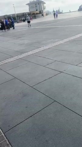The video shows a pedestrian area with a building that appears to be a restaurant or bar with outdoor seating. The building has a green awning and an American flag is flying from a flagpole. Several people are visible walking and sitting at tables. Bicycles are parked outside the establishment. The ground is paved with concrete and some brickwork. The sky is cloudy.