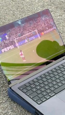 A close-up shot shows a laptop screen displaying a soccer penalty kick in progress. A player in a white and red jersey is about to kick the ball toward the goal defended by a goalkeeper in green. The background is filled with a large crowd of spectators. The laptop is resting on a blue cushion on an asphalt surface. The laptop's keyboard and touchpad are visible. The camera then shifts to the reaction of a group of men watching the game. Two men are visible: one is wearing a white hoodie, and the other is in a black t-shirt. They are watching intently, and the man in the black t-shirt puts his hands on his head in disbelief. The background is slightly blurred, suggesting they are outdoors. The sound includes crowd noise and the excited commentary of a soccer game.