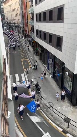 The video shows a street procession from a high-angle perspective. The street is lined with buildings and features a pedestrian crossing with white stripes. A large group of people, mostly wearing white shirts and dark pants or skirts, are marching in formation down the street. They appear to be a band or a marching group, carrying flags and possibly instruments. Several individuals, predominantly young women, are seen in the foreground, also holding flags, some of which are colorful and bear logos. They are positioned closer to the camera, on a sidewalk area that is separated from the street by a metal railing. A few spectators are visible on the balconies of the buildings, observing the procession. The street is paved and has yellow markings, including a zigzag pattern and a disabled parking symbol. There are also parked motorcycles and a stroller on the sidewalk. The overall scene suggests a public event or celebration taking place in an urban environment.