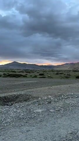 This is a vertical, handheld video taken from the perspective of someone looking out over a wide, desolate landscape at dusk. The foreground is a rough, rocky surface, possibly a dirt road or an unpaved area. Further back, there is a flat, sparsely vegetated plain covered in low-lying bushes. In the distance, a range of mountains stretches across the horizon. The sky is filled with dramatic, dark, and cloudy formations, with a sliver of orange and yellow light from the setting sun breaking through the clouds in the center of the frame, illuminating the distant peaks. The lighting suggests a cool, twilight atmosphere. There are no people or obvious man-made structures visible. The video appears to be a static shot with no significant movement other than very slight camera shake.