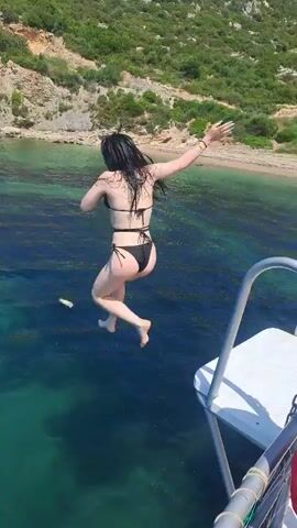 A person, likely a woman, wearing a black bikini, jumps into clear turquoise and blue water from what appears to be a boat or platform. A white object floats on the surface of the water further out. A large splash is visible where the person enters the water. In the background, a rocky coastline with some green vegetation and a small sandy beach is visible under bright daylight conditions. Part of a boat's railing and netting are visible on the right side of the frame, indicating the jump is from a vessel. The water is calm and transparent, revealing the seabed in shallower areas. The scene depicts a leisure activity on a sunny day in a coastal environment, possibly near Selçuk, Turkey, as per the provided context.