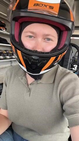 A young woman in a helmet and polo shirt is the subject of a selfie-style video. She is seated in a go-kart, wearing a black, orange, and white helmet with the brand name "BOX'S" on it. The helmet's interior is pink. She also wears a black head covering beneath the helmet and a light olive green polo shirt. Below her shirt, blue jeans are visible.

Her face, partially obscured by the helmet's visor, reveals light blue eyes and a smile. She gives a hand gesture resembling the "rock on" sign. Her nails are painted a light lavender.

The background shows a go-kart garage with other go-karts parked nearby on shelves. The go-kart she's in has "SOOS" printed on the side. The video's perspective is from the person's point of view, suggesting it was taken with a handheld device.
